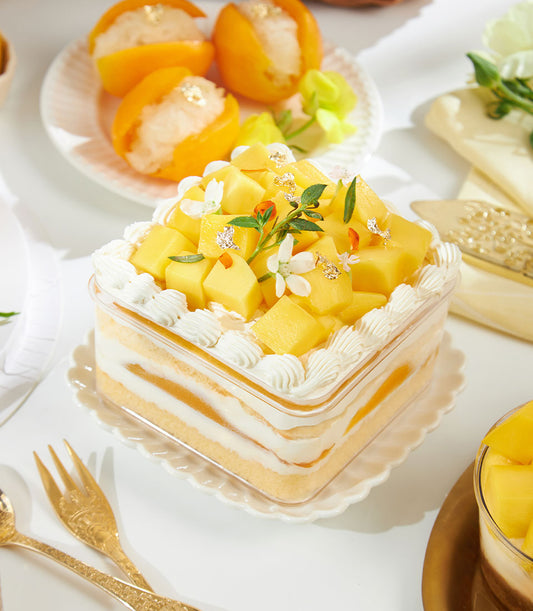 Layered mango cream cake with fresh fruit, white flowers and gold leaf in clear container