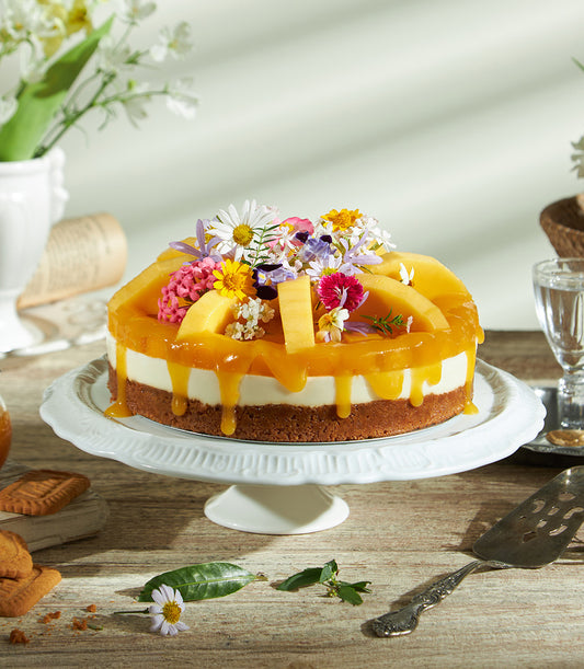 Tropical mango cheesecake with creamy layers, vibrant glaze, fresh fruit, and edible flowers on ceramic stand