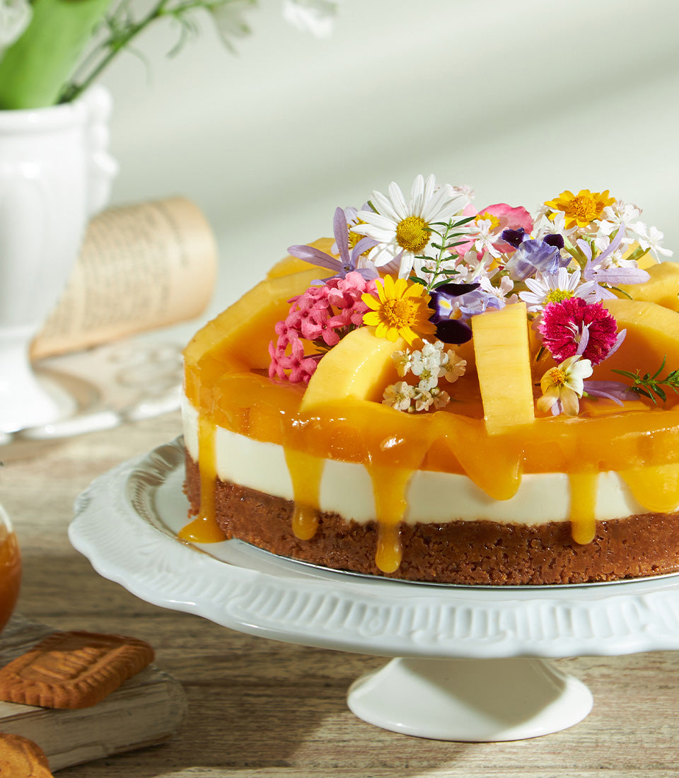 Mango cheesecake with orange glaze, fresh flowers, and tropical fruit on ceramic stand