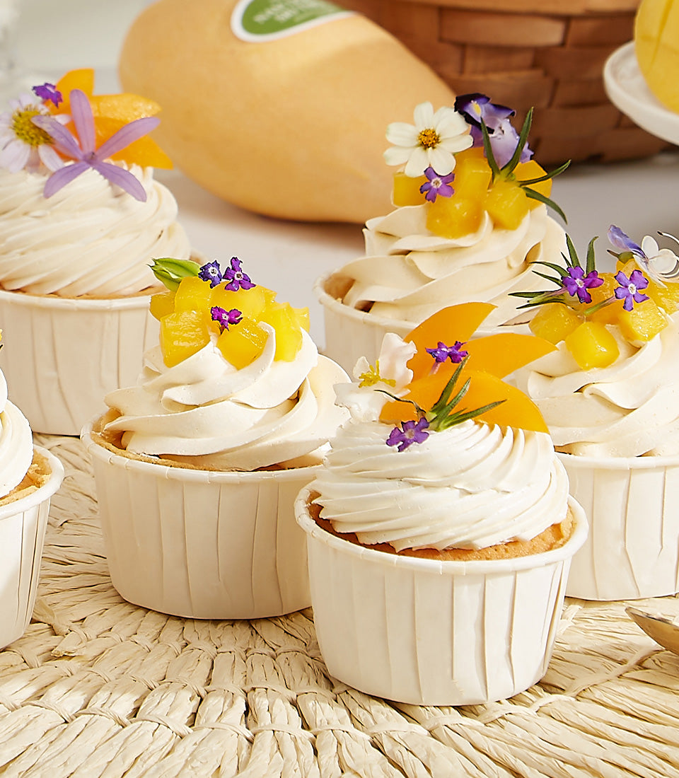 Mango cupcakes with fresh fruit, white frosting, and purple flowers on woven mat