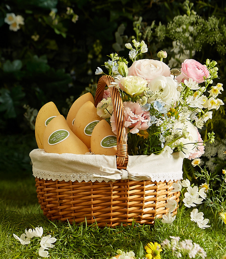 Rustic wicker gift basket with golden Nam Dok Mai mangoes, pink ranunculus, and white flowers on lace-lined fabric