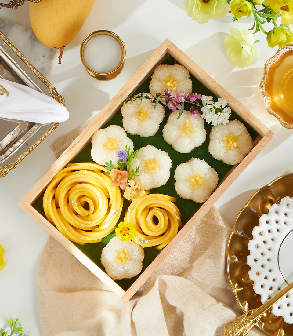 Mango sticky rice dessert box with eight portions, mango slices, edible silver leaf, and fresh flowers on banana leaf in