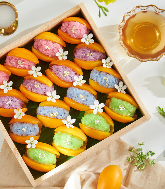 Vibrant mango sticky rice gift box with pastel rainbow rice, gold leaf, and mango halves on banana leaf
