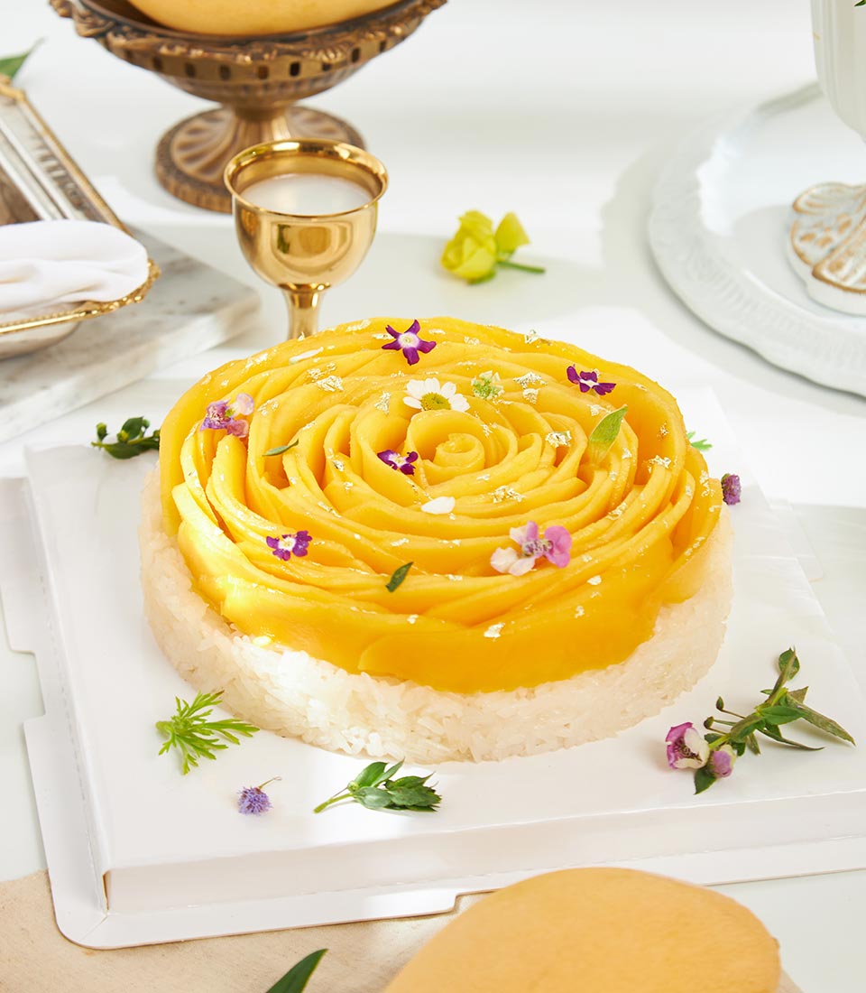 Golden rose mango sticky rice cake with edible gold leaf and purple flowers on white board