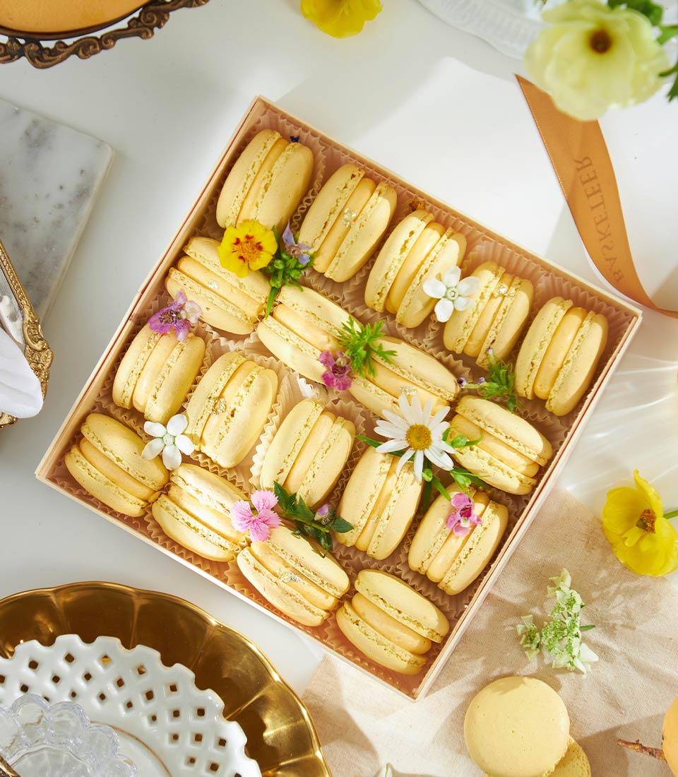 Golden mango macarons with fresh flowers and silver leaf accents in luxury gift box