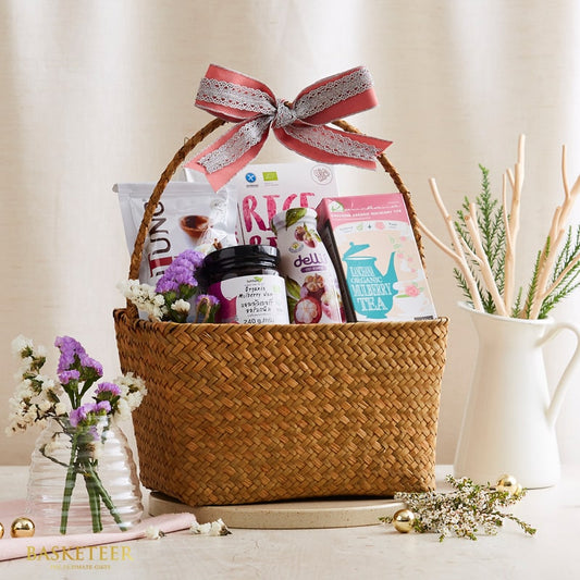 Woven straw gift basket tied with coral-pink ribbon, filled with organic mulberry jam, tea, and wellness products