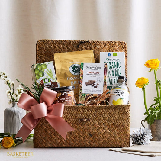 Woven straw gift basket with pink ribbon bow filled with gourmet coffee, chocolates, crackers, and artisan treats