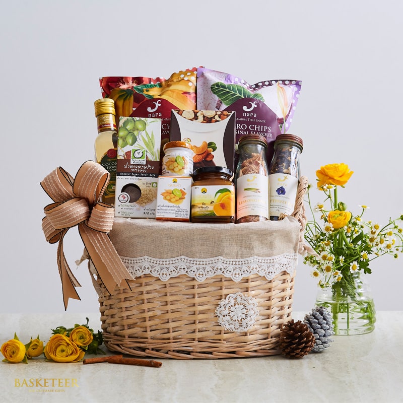 Wicker gift basket with tropical snacks, nuts, and gourmet items, adorned with white lace and brown ribbon bow, alongside