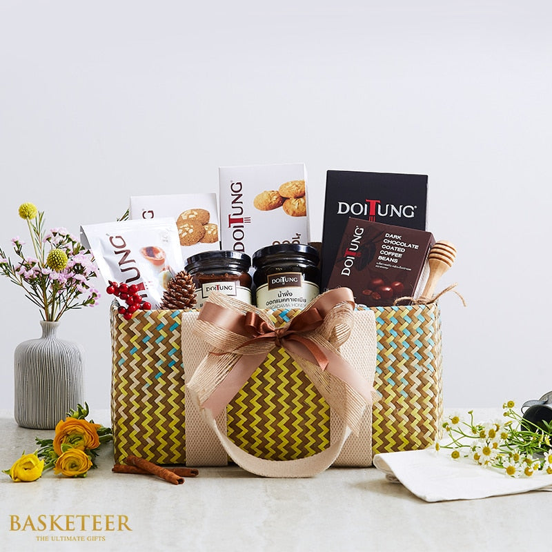 Woven gift basket with Thai gourmet treats, coffee, chocolate, and honey dipper, styled with pink and yellow flowers