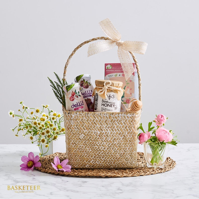 Woven rattan gift basket with honey, gourmet beverages, tea, and fresh daisies and peonies