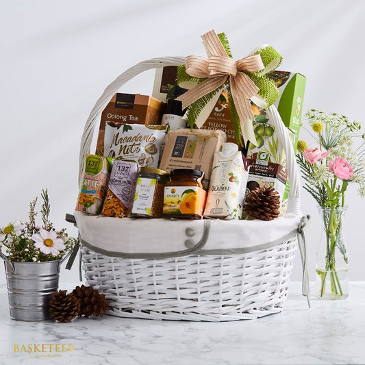 White wicker gift basket with Thai gourmet foods, tea, nuts, and preserves, tied with peach and green ribbon bow