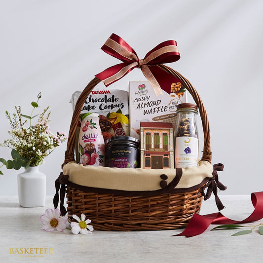 Woven brown wicker gift basket with red gold-trimmed ribbon bow, filled with gourmet Thai treats and artisan snacks, paired