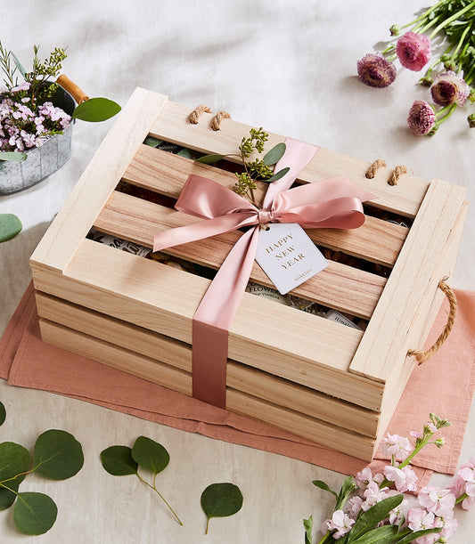 Wooden gift crate filled with gourmet wellness products, tied with dusty rose ribbon, surrounded by fresh pink flowers and