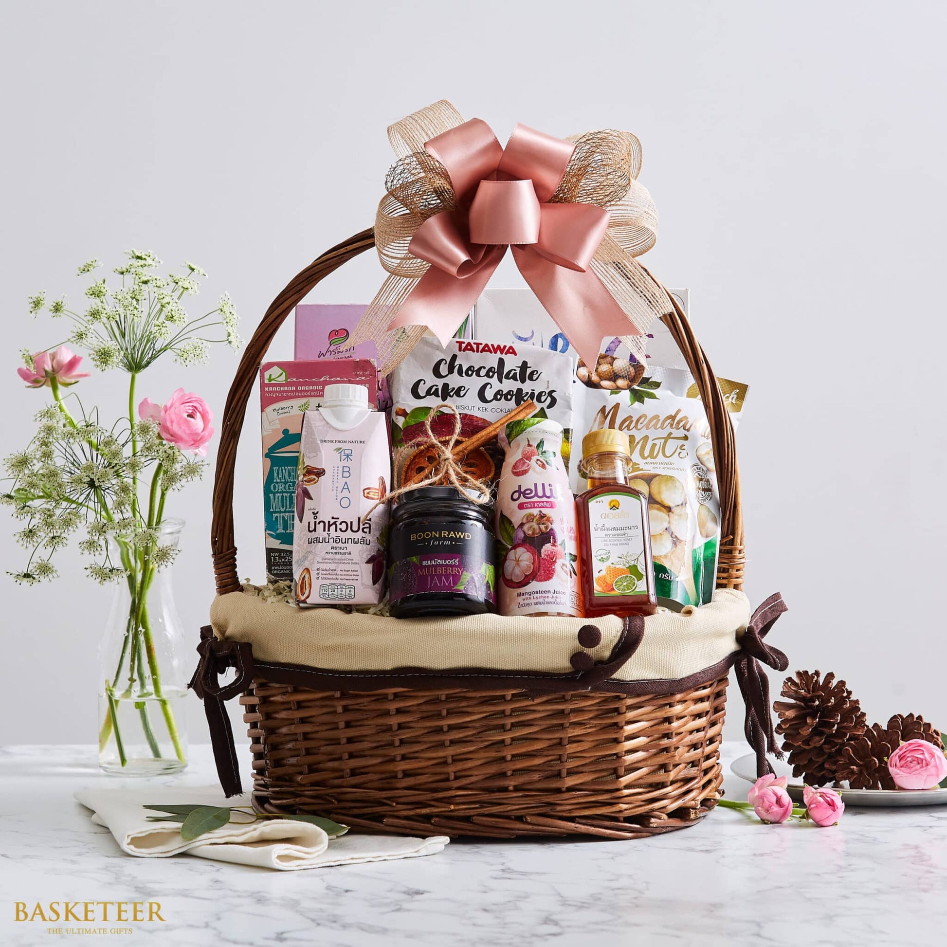 Large woven wicker gift basket with rose gold satin bow, filled with Thai gourmet foods, beverages, and treats on marble