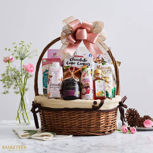 Large woven wicker gift basket with rose gold satin bow, filled with Thai gourmet foods, beverages, and treats on marble