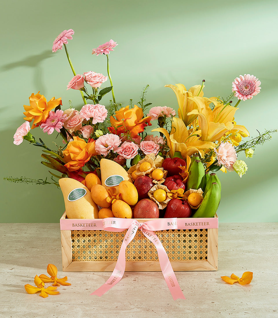 Tropical fruit gift basket with yellow mangoes, apples, bananas and pink, orange, yellow flowers in woven rattan basket