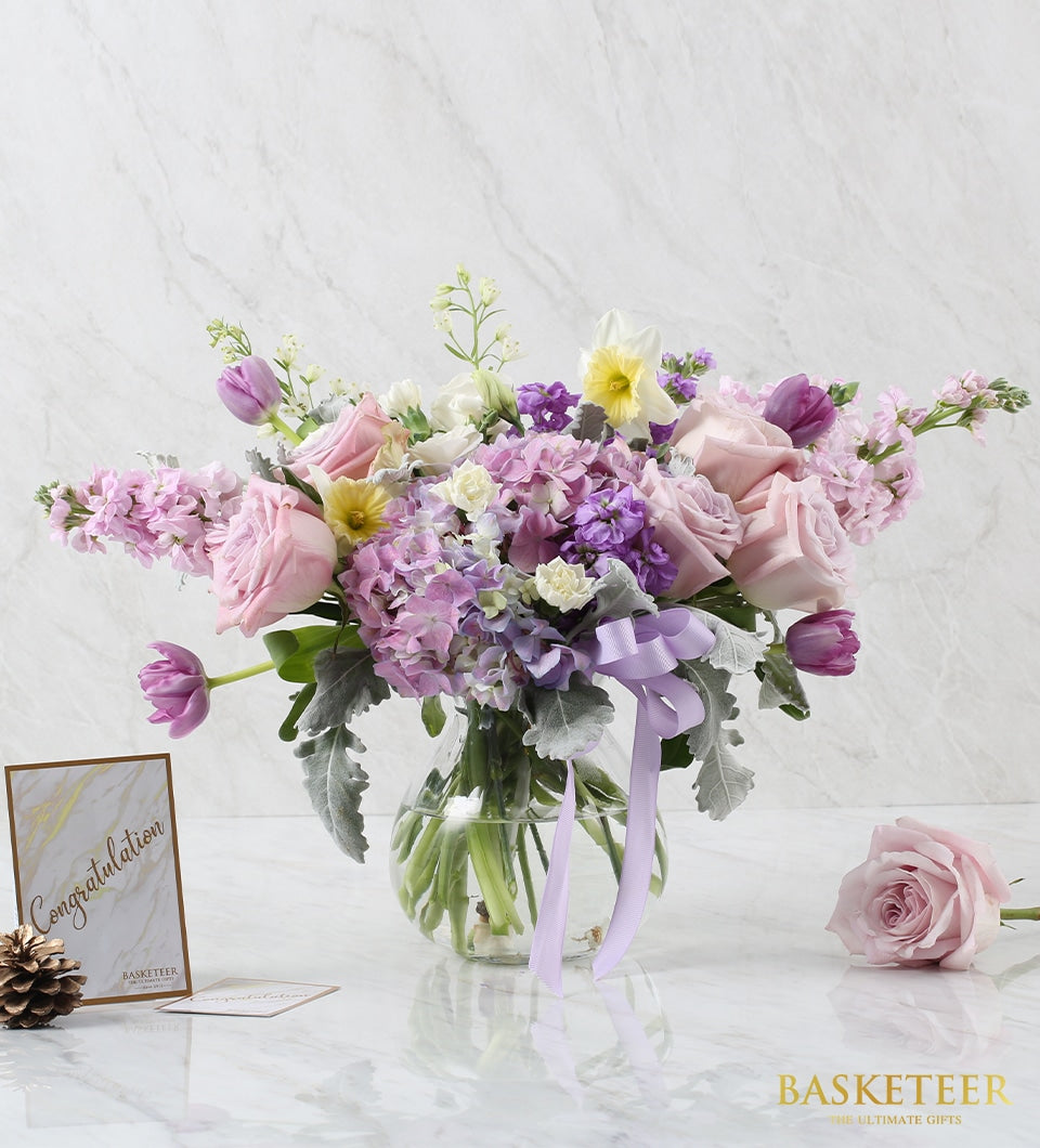 Vibrant purple and pink mixed flower arrangement in clear glass vase with congratulations card
