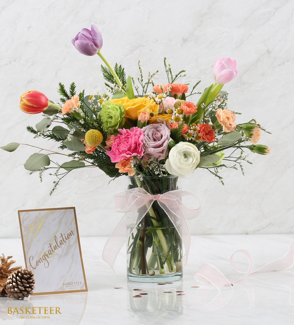 Vibrant mixed flower arrangement in clear glass vase with tulips, roses, and carnations, tied with pink ribbon