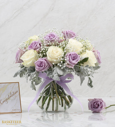 Purple and cream roses with baby's breath in clear glass vase, tied with lavender ribbon