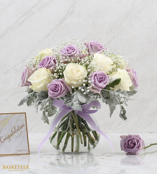 Purple and cream roses with baby's breath in clear glass vase, tied with lavender ribbon
