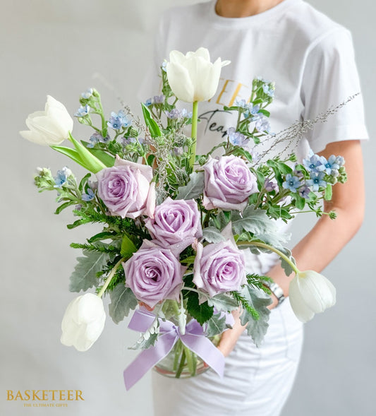 Purple roses and white tulips with dusty miller in clear glass vase, tied with lavender ribbon