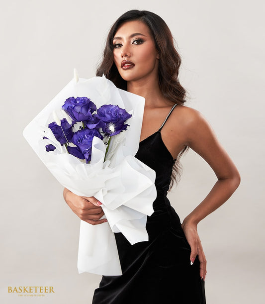 Deep purple Eustoma bouquet with dried botanicals in white tissue wrap, held by woman in black velvet dress