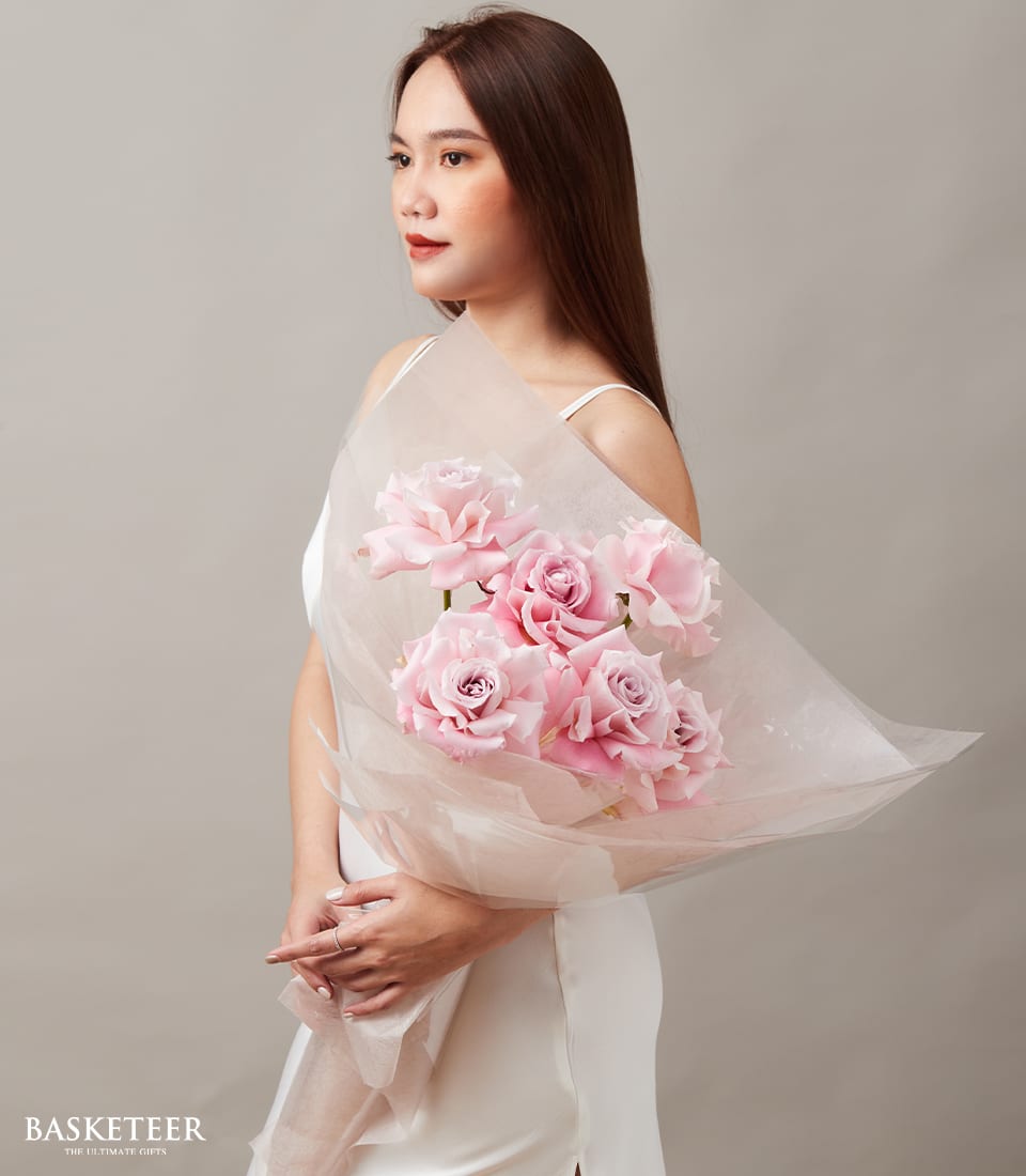Blush pink rose bouquet with six blooms in soft tissue wrap, held by woman in white dress