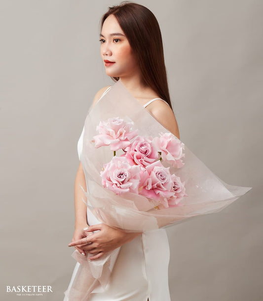 Blush pink rose bouquet with six blooms in soft tissue wrap, held by woman in white dress