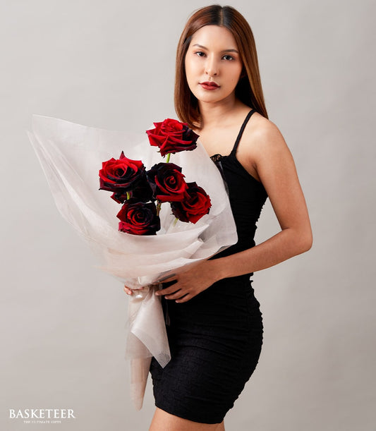 Red and black-centered roses bouquet wrapped in tissue, held against neutral background for Valentine's Day gifting