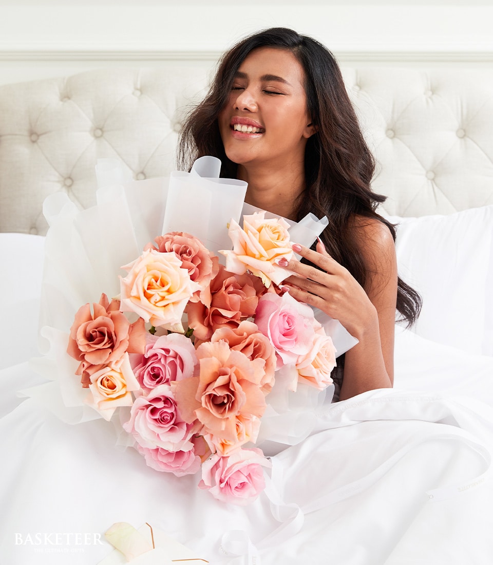 Woman in bed smiling with large bouquet of pink, peach, and orange roses wrapped in white paper