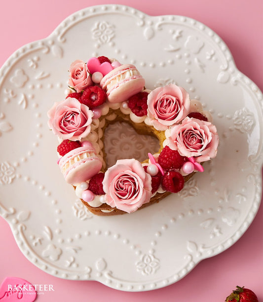 Heart-shaped vanilla cake with fresh pink roses, raspberries, macarons, and sugar pearls on floral ceramic plate