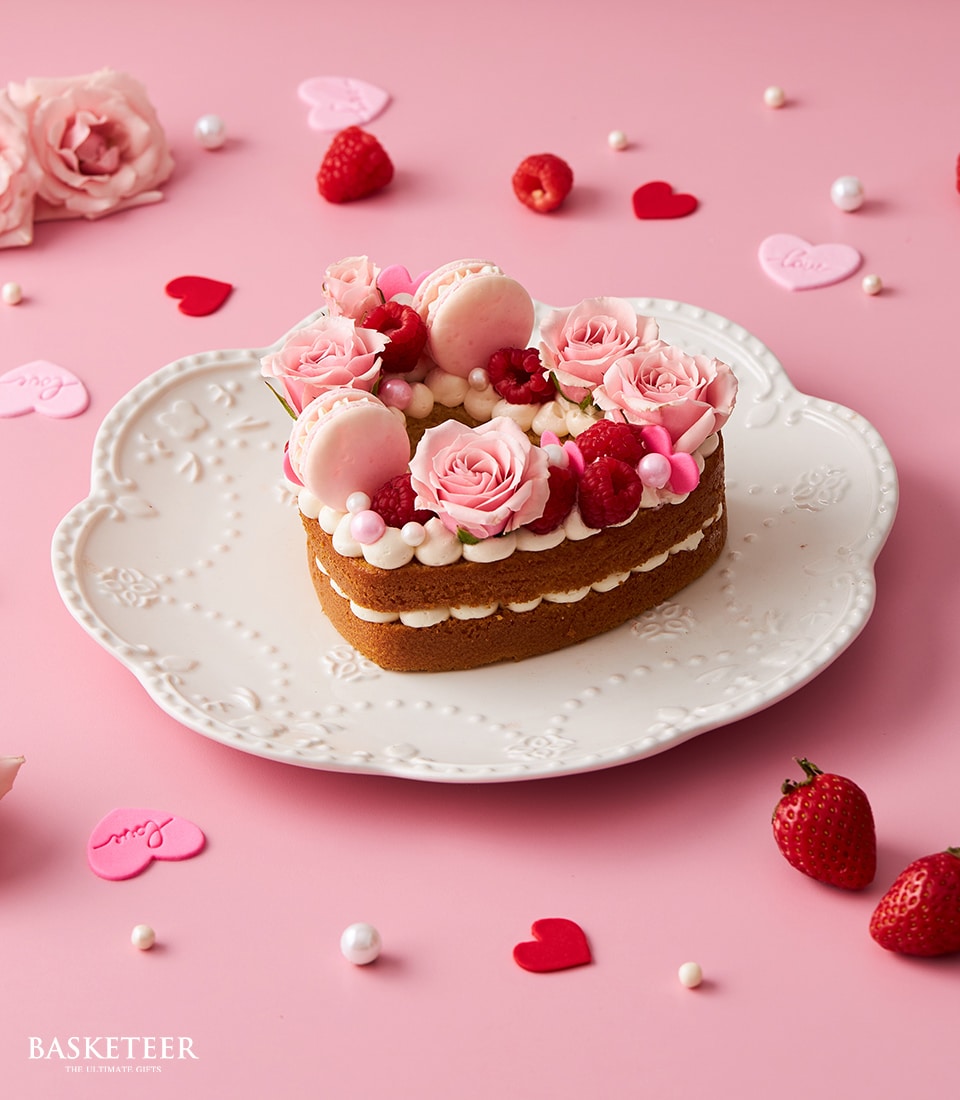 Heart-shaped vanilla cake with pink roses, macarons, and fresh raspberries on white scalloped plate