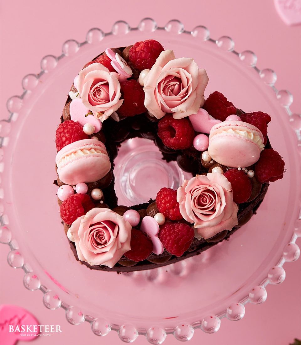 Heart-shaped dark chocolate cake with raspberries, roses, and macarons on glass stand