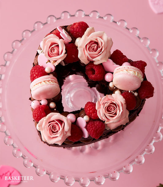 Heart-shaped dark chocolate cake with raspberries, roses, and macarons on glass stand