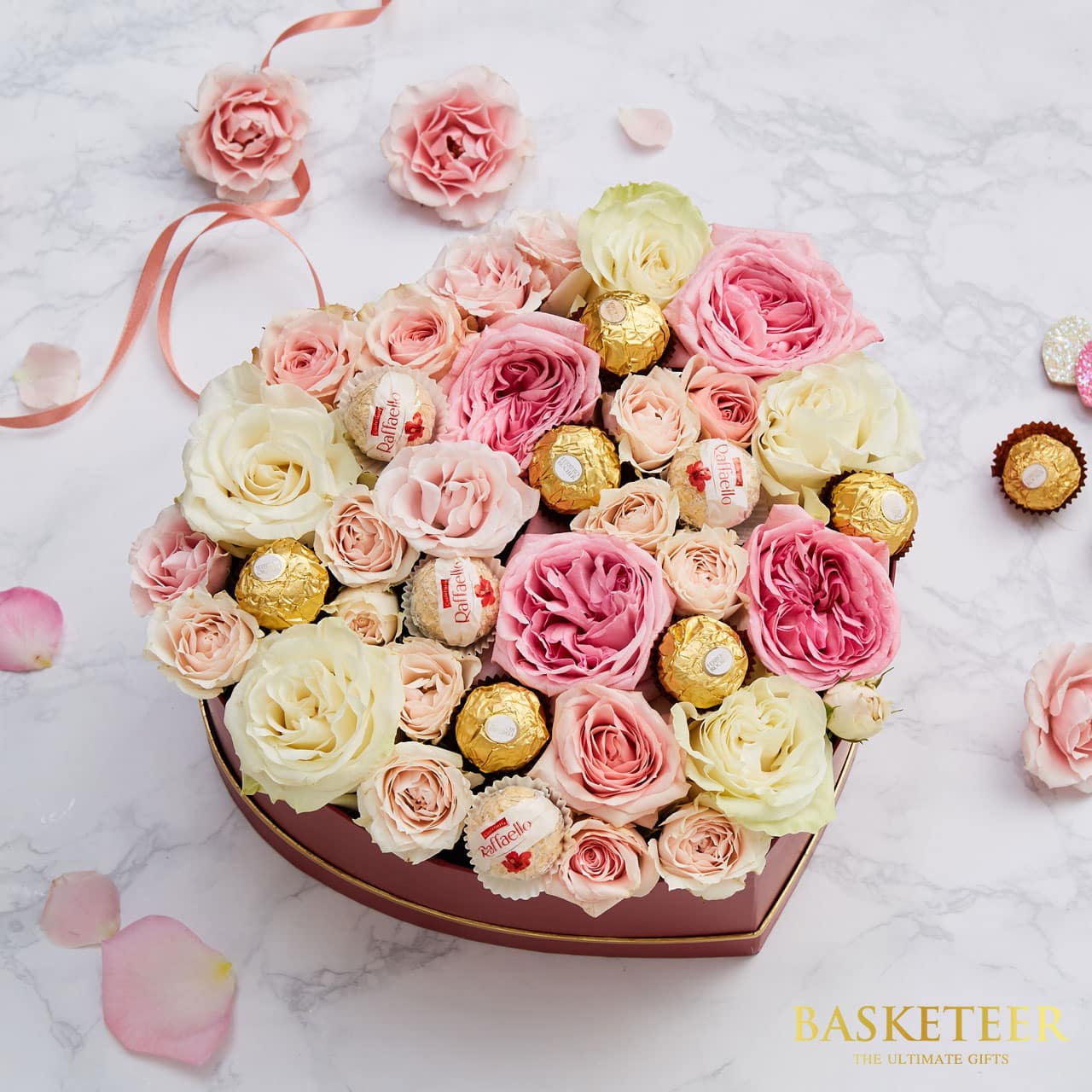 Heart-shaped burgundy gift box with pink and cream roses, gold-wrapped chocolates, and romantic rose petals for anniversary