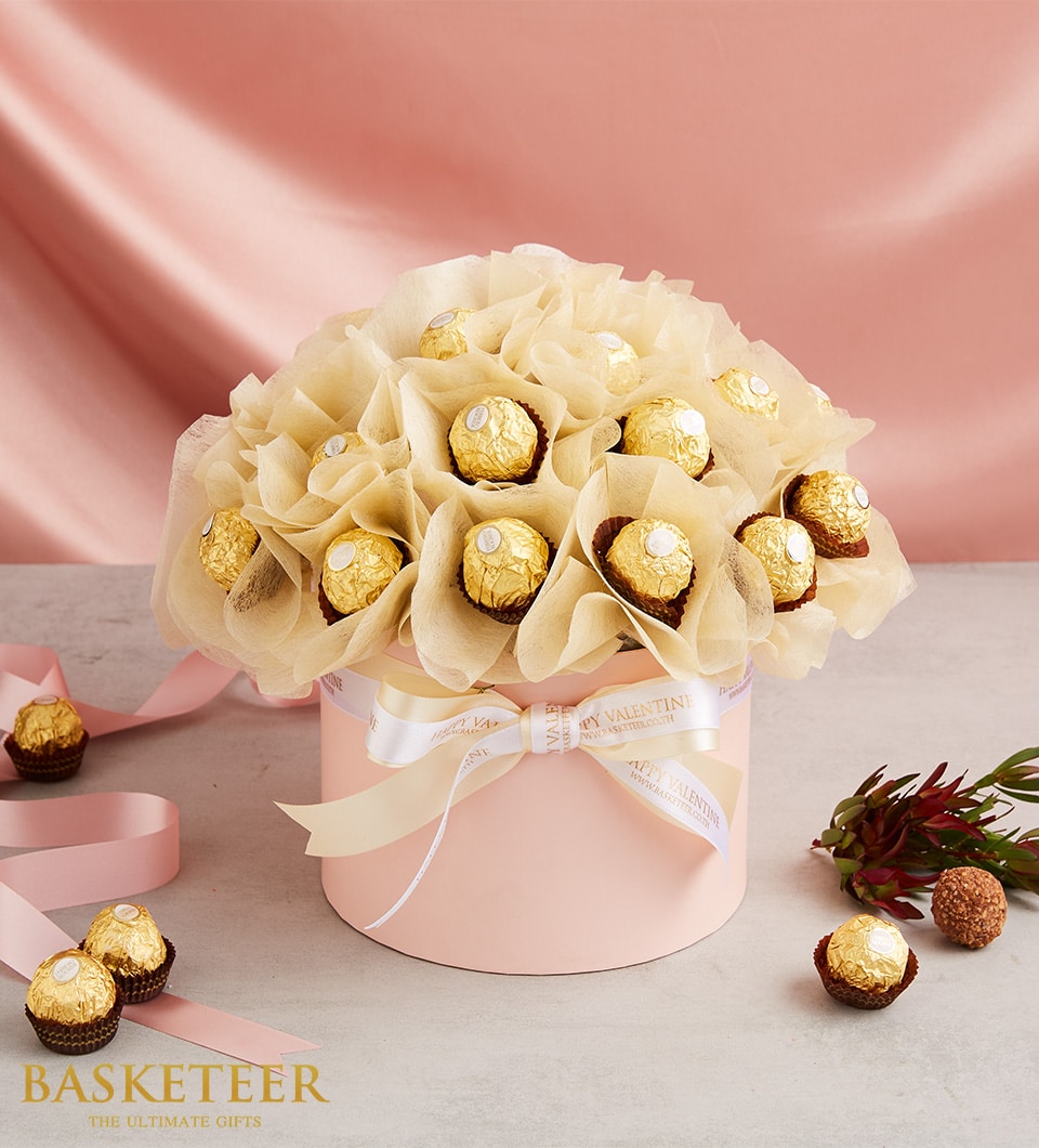 Gold-wrapped Ferrero Rocher chocolates in pink gift box with cream tissue, Valentine's Day ribbon, and romantic foliage