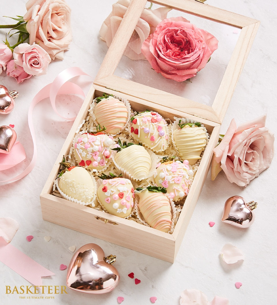 White chocolate-covered strawberries with pink drizzle and sprinkles in wooden gift box, surrounded by roses and rose gold