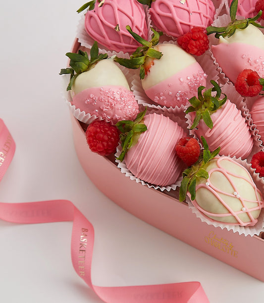 Heart-shaped box of artisan chocolate-covered strawberries with pink and white chocolate, gold details, and fresh raspberries