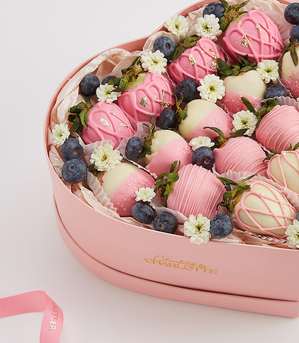 Heart-shaped box of chocolate-covered strawberries with pink and white coating, gold leaf, and fresh blueberries for