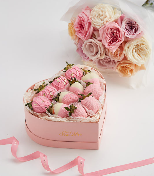 Heart-shaped box of pink and white chocolate-covered strawberries with gold accents, paired with fresh pink and cream roses