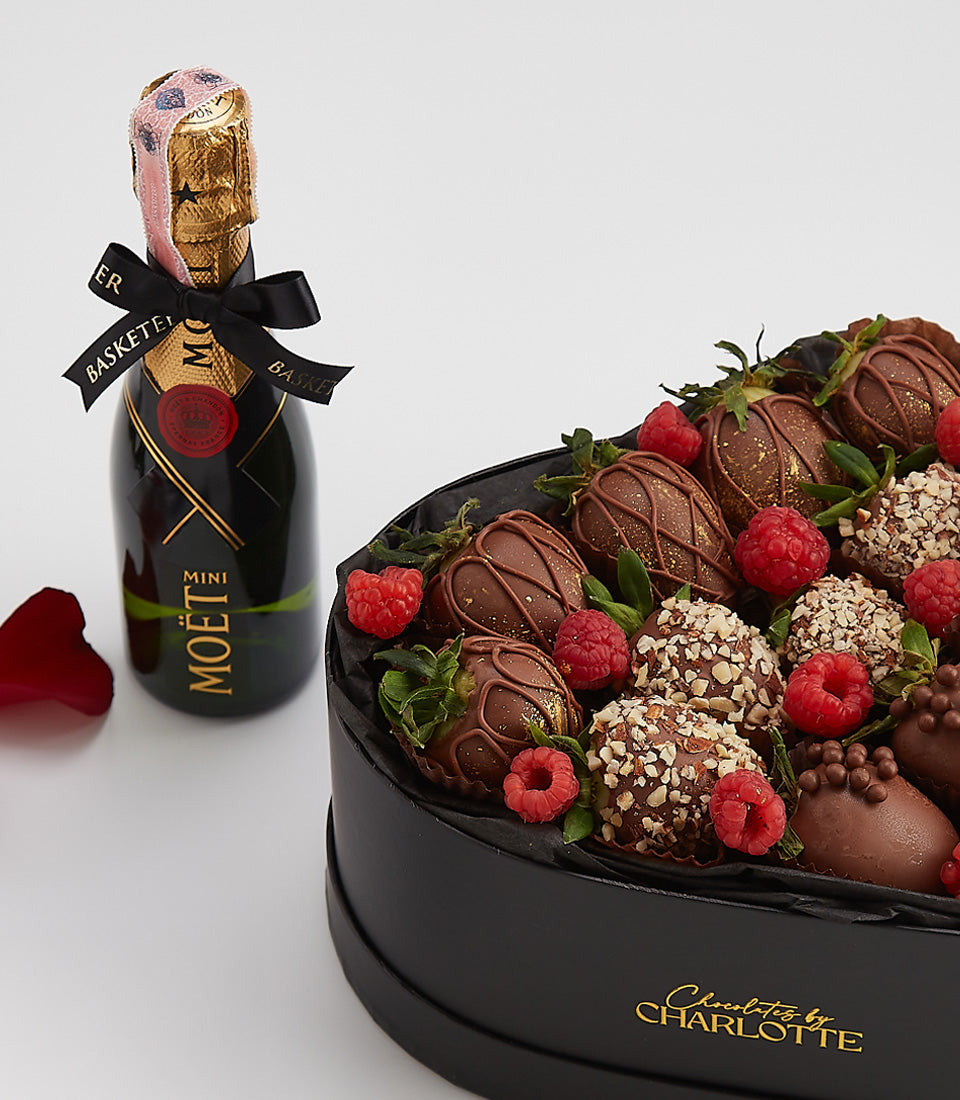 Heart-shaped black box of dark chocolate strawberries with gold drizzle, mini Moët champagne, and fresh raspberries for