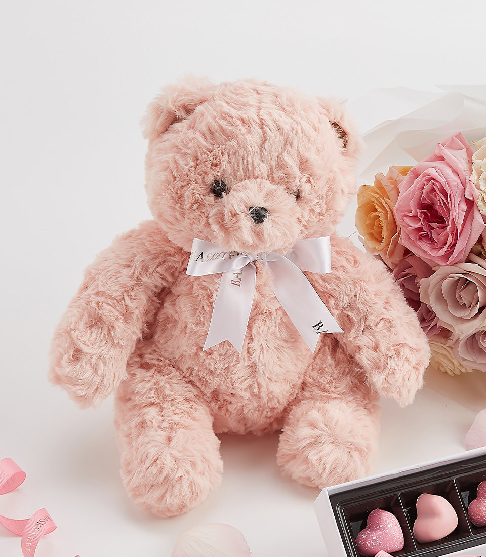 Pink teddy bear with white satin bow, fresh roses in blush and peach tones, and heart-shaped chocolates Valentine's gift