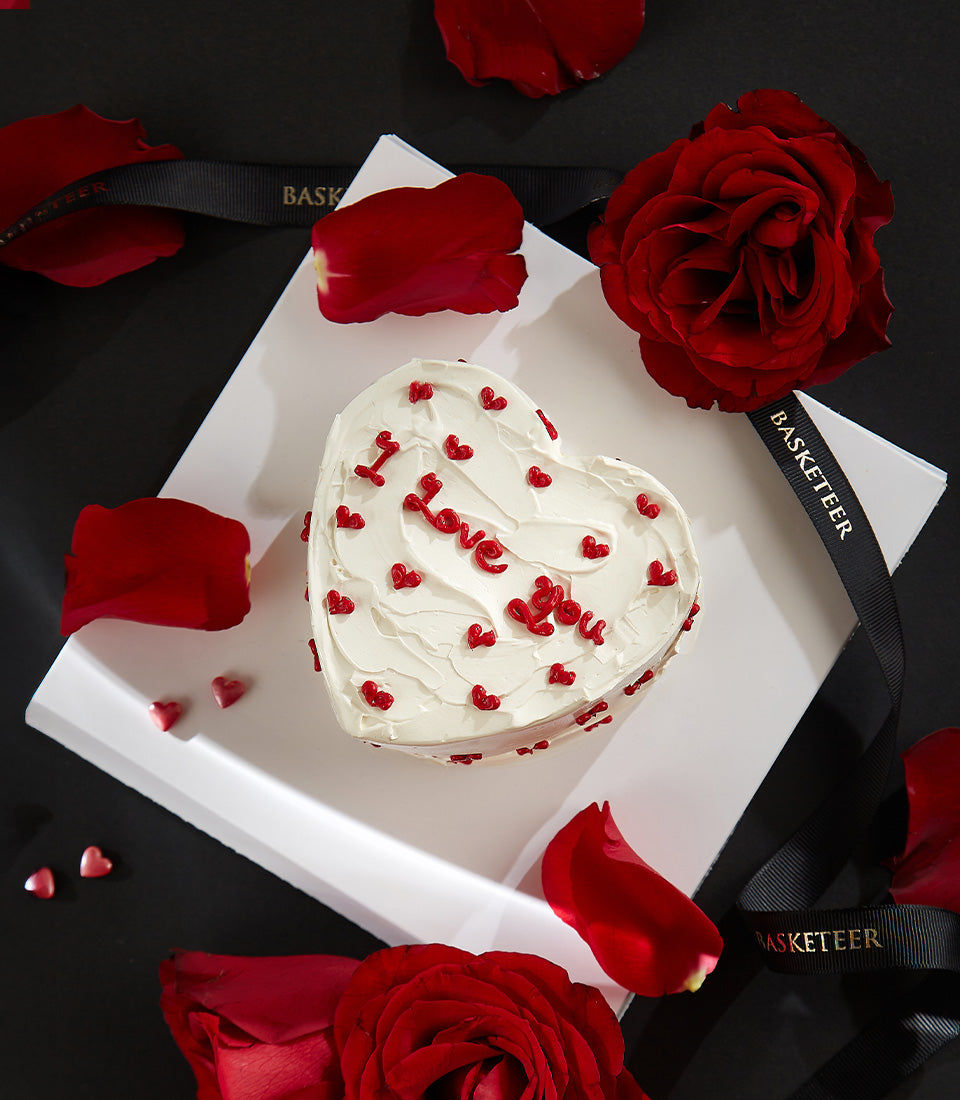 Heart-shaped chocolate raspberry cake with white frosting, red "I Love You" lettering, surrounded by red roses and rose