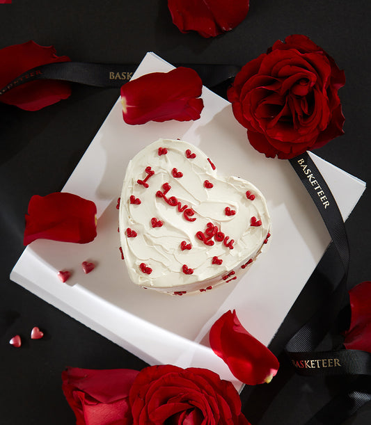 Heart-shaped chocolate raspberry cake with white frosting, red "I Love You" lettering, surrounded by red roses and rose
