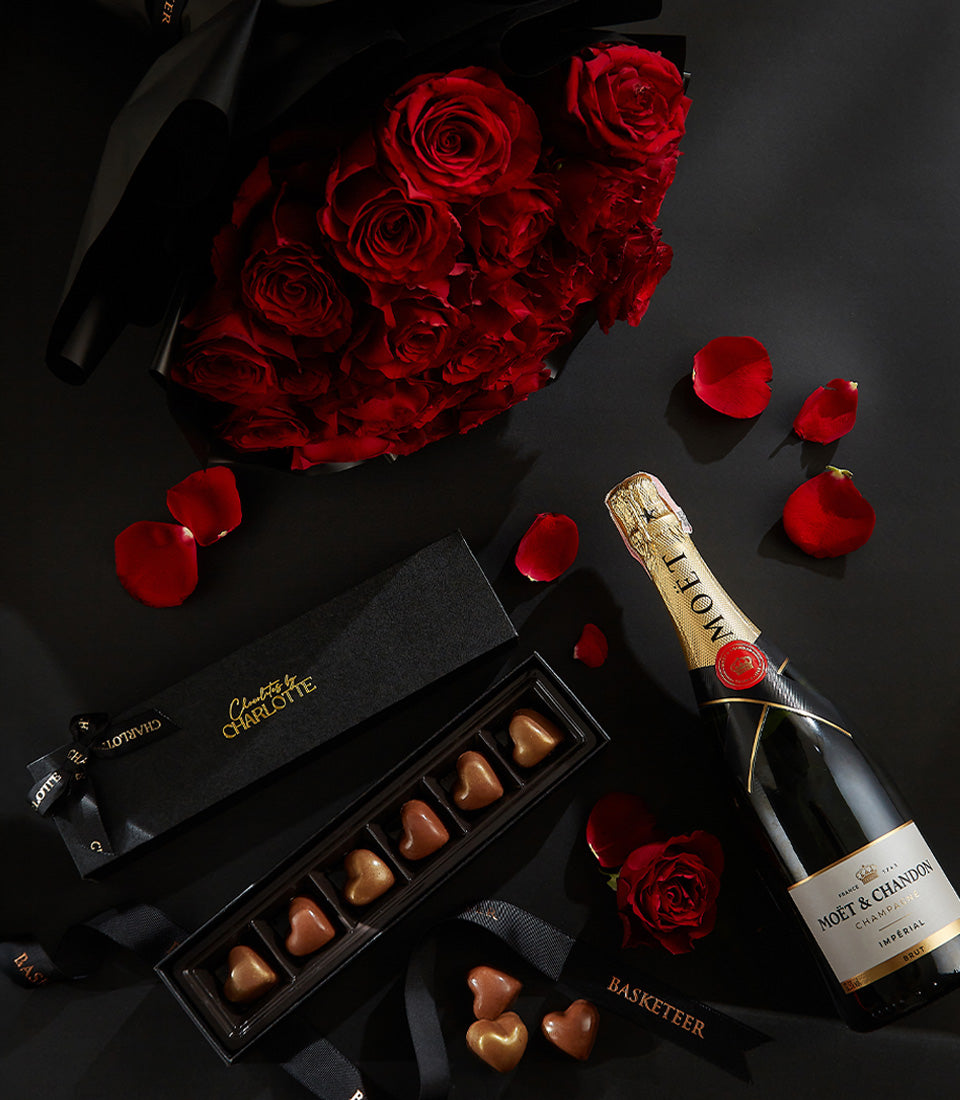 Luxury Valentine's champagne gift set with deep red roses, heart-shaped chocolates, and gold-foil bottle on dark surface