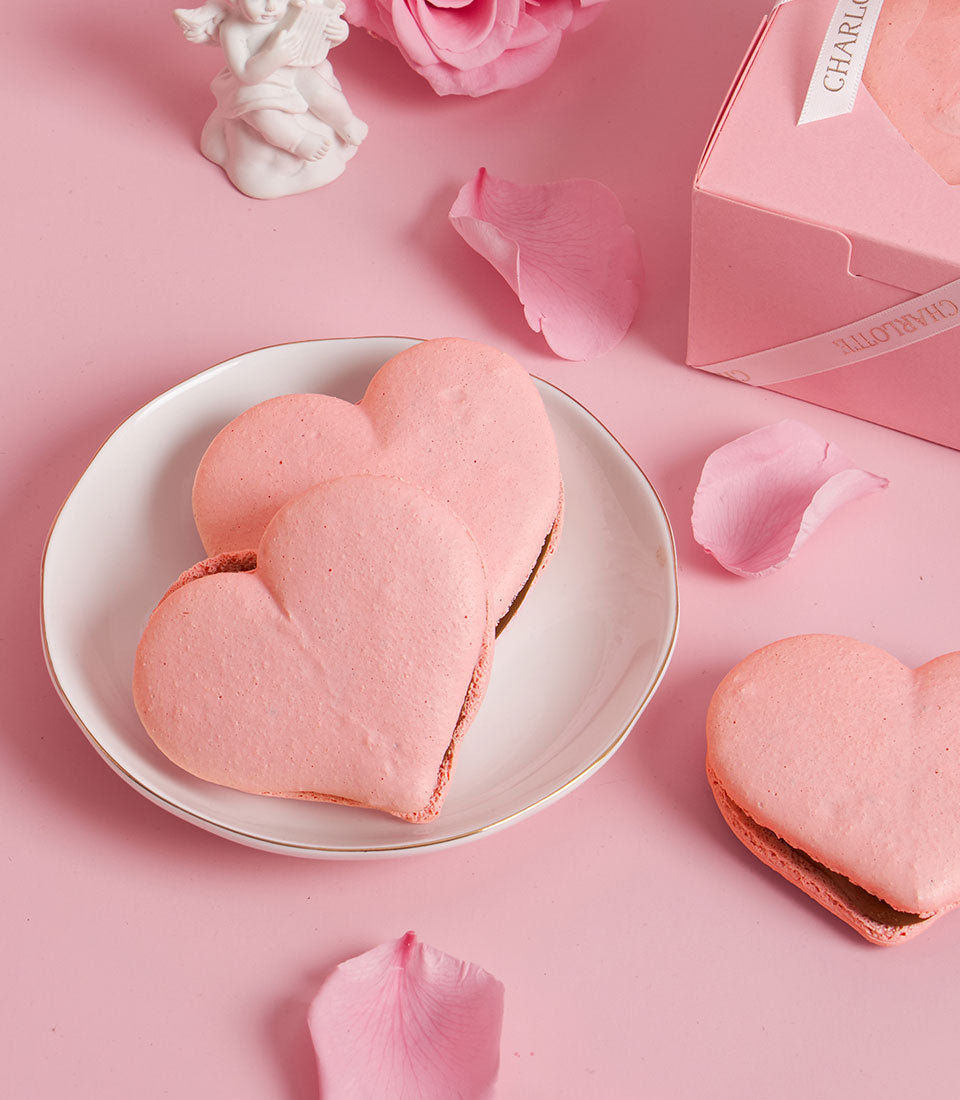 Charlotte Bakery artisan heart-shaped cookies, a romantic pastry gift. Pink macarons sit on a plate with rose petals and a bakery gift box. Perfect custom cake Bangkok treat.