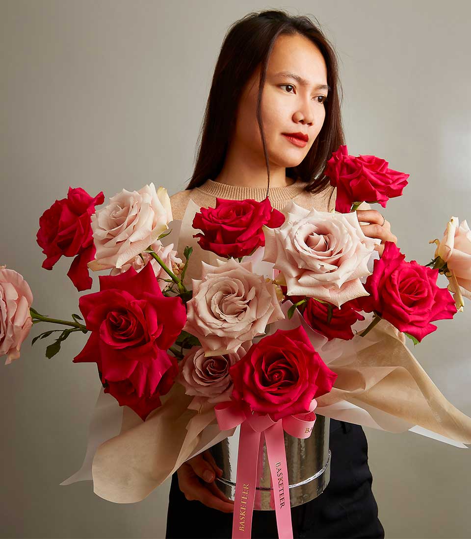 Romantic pink and red roses in silver cylinder with brown paper wrap and pink ribbon, held by woman in cream knit top