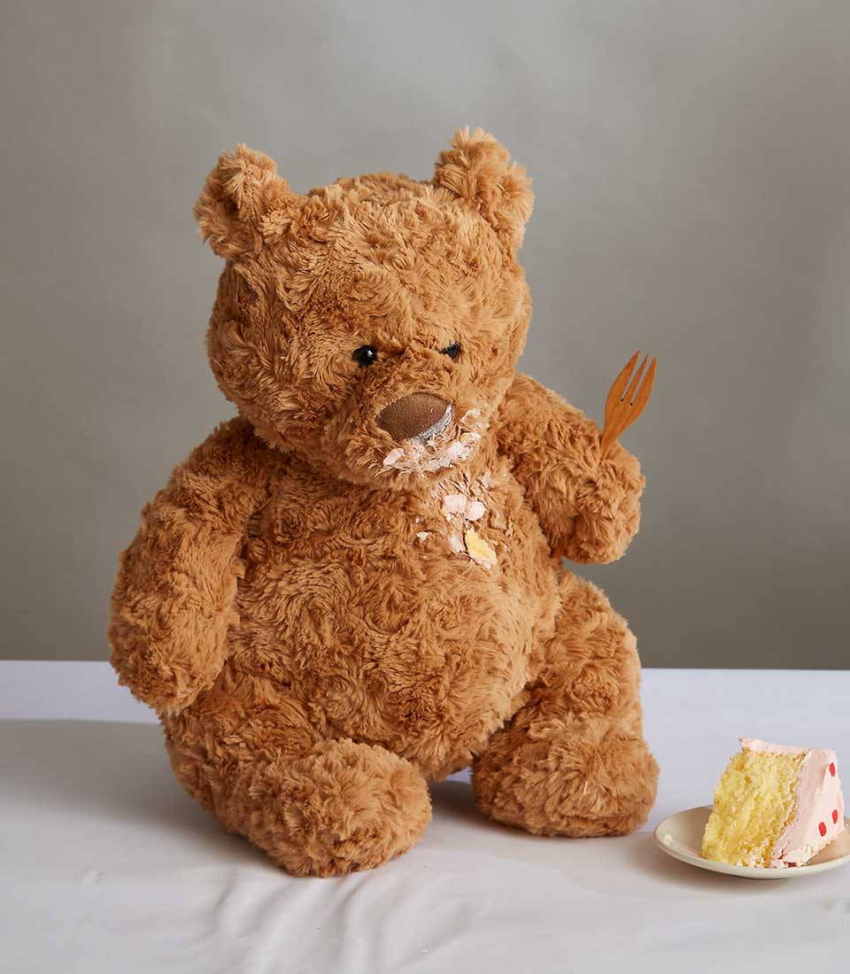 Plush teddy bear with frosting-smeared face holding wooden fork beside slice of yellow cake with pink frosting