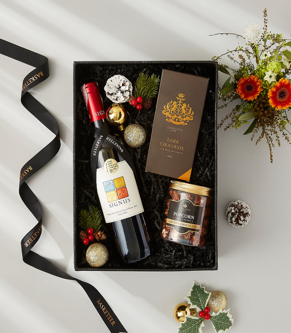 Luxury holiday gift box with red wine, dark chocolate, and gourmet popcorn, dressed in gold and festive greenery