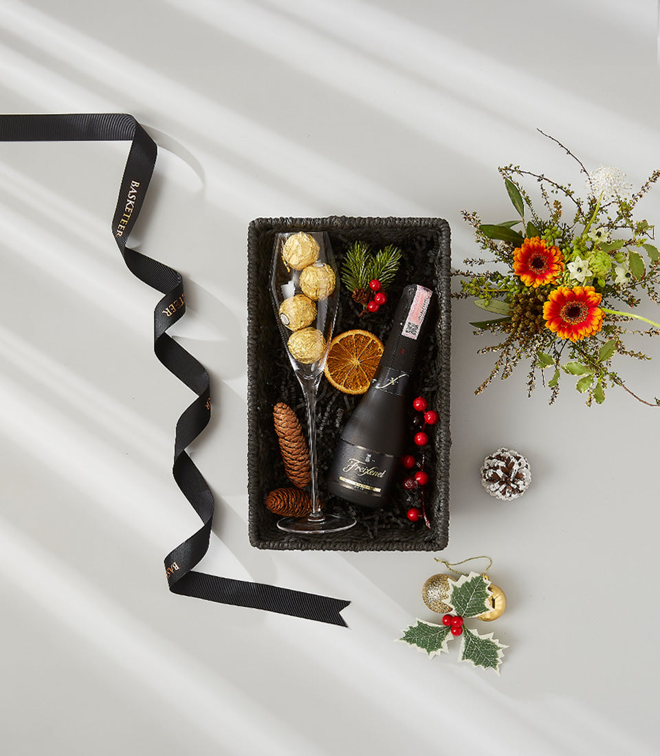 Festive black woven gift basket with champagne flute, Freixenet sparkling wine, gold chocolates, and holiday greenery